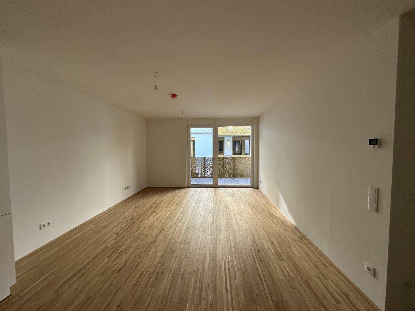 Geräumiger Wohnbereich mit hellem Parkettboden und Zugang zu einem Balkon.