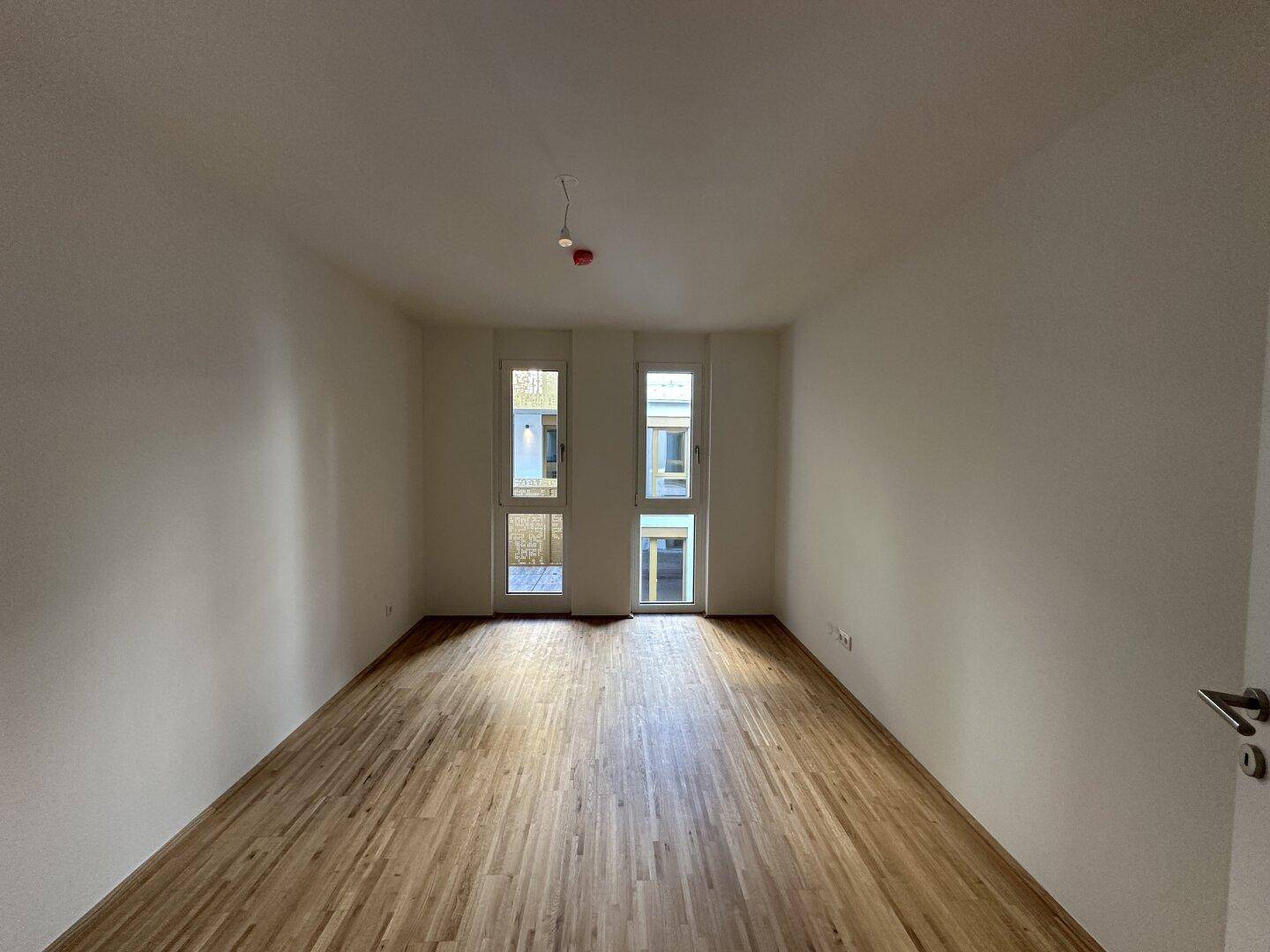 Helles Zimmer mit Parkettboden und zwei Fenstern, die viel Tageslicht hereinlassen.