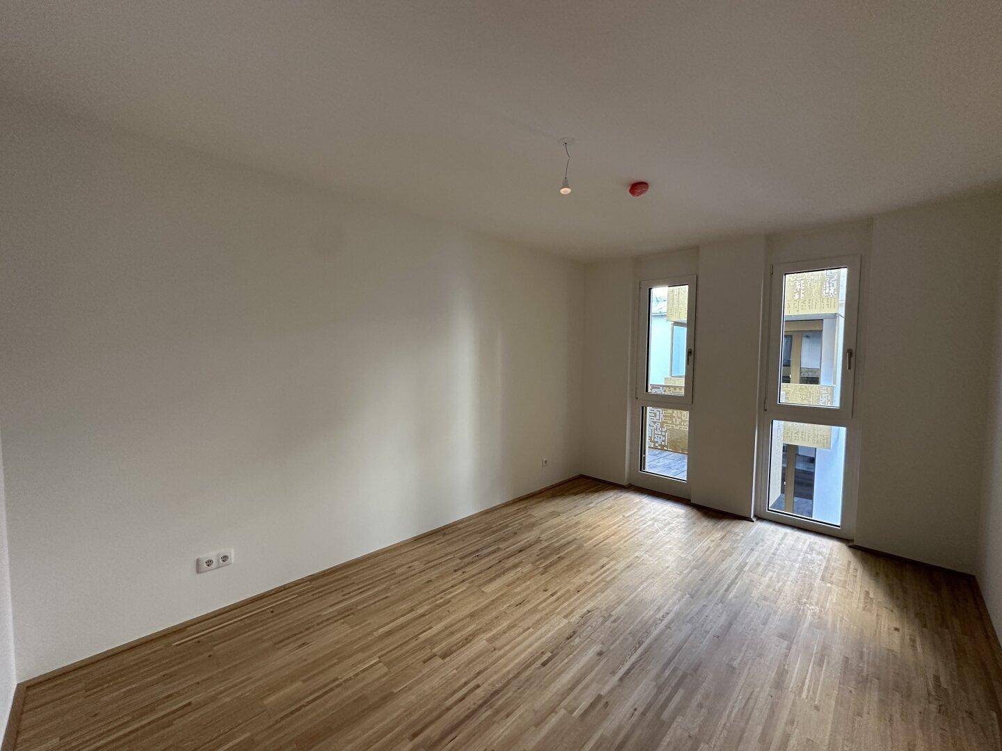 Helles Zimmer mit Parkettboden und zwei Fenstern, die einen Blick nach draußen bieten.