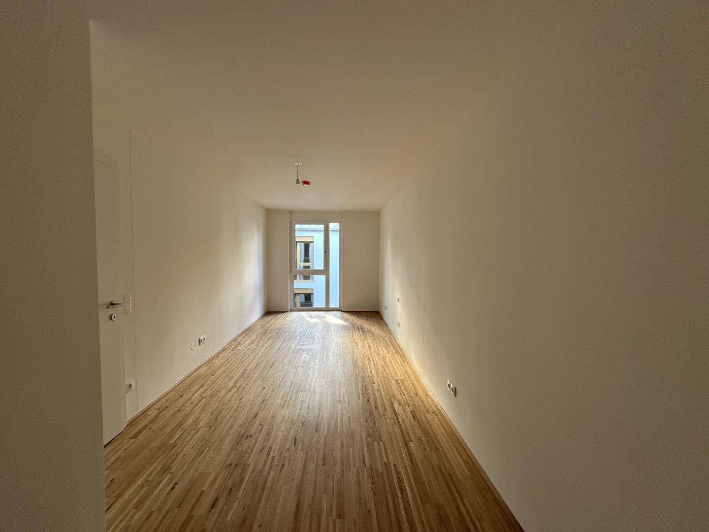 Langes, helles Zimmer mit Parkettboden und einer Tür, die zu einem Balkon führt.