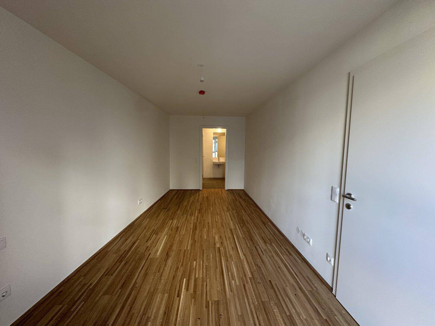 Leeres Zimmer mit Holzboden und Blick auf das angrenzende Badezimmer, ideal als Schlafzimmer.
