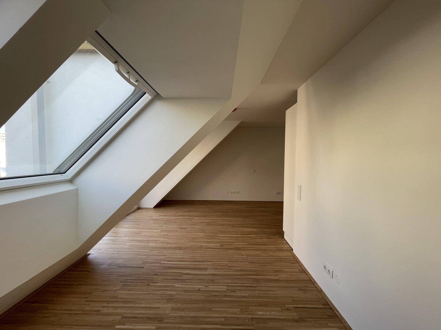 Helles Zimmer im Dachgeschoss mit Holzboden, schrägen Wänden und einem großen Dachfenster für viel Tageslicht.