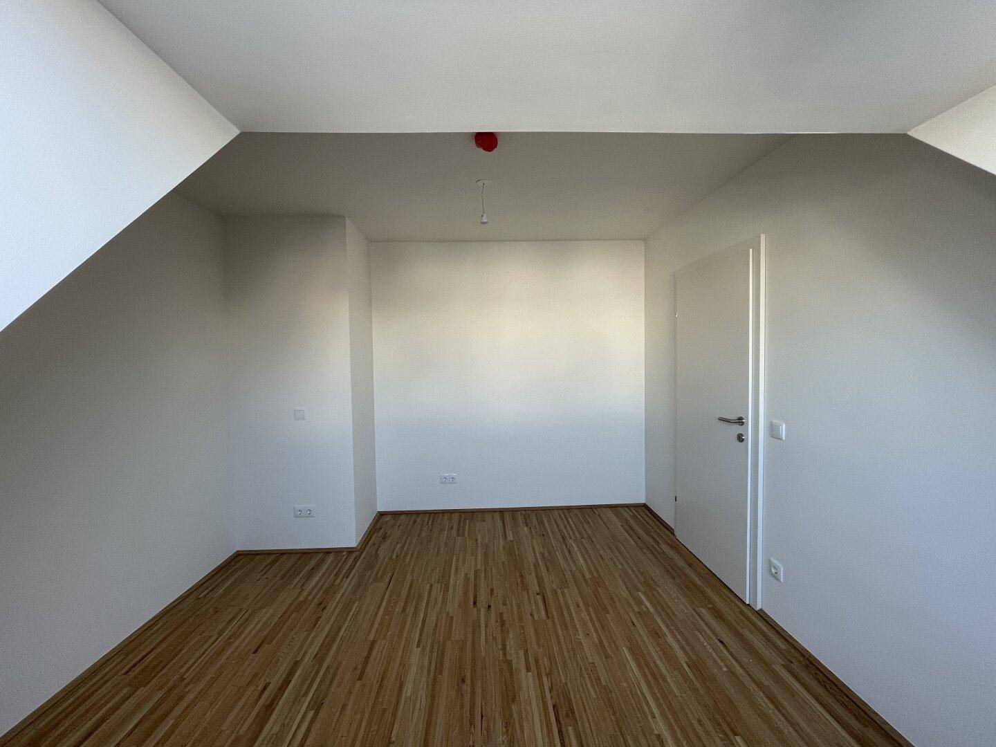 Geräumiges Zimmer im Dachgeschoss mit Parkettboden, Dachschräge und weißer Tür.