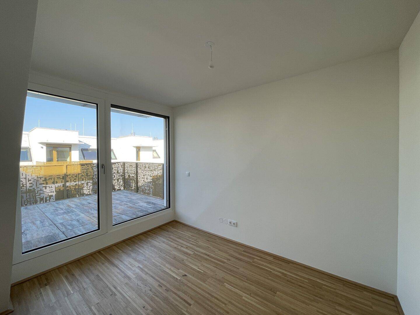 Heller Raum mit Holzboden und großen Fenstern, die auf eine Terrasse führen.