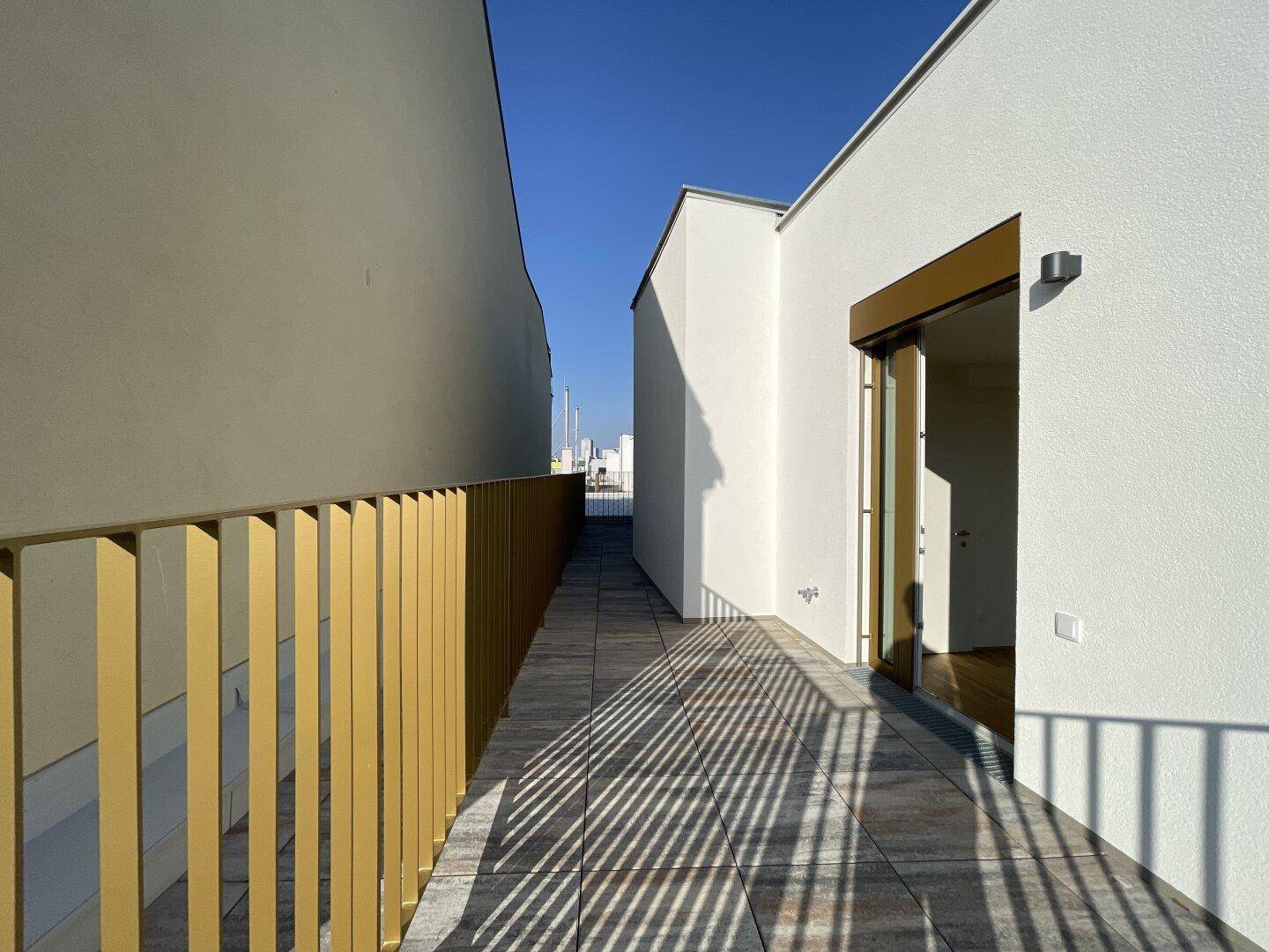 Schmaler Terrassenbereich mit Fliesenboden und goldener Brüstung zwischen Gebäuden.