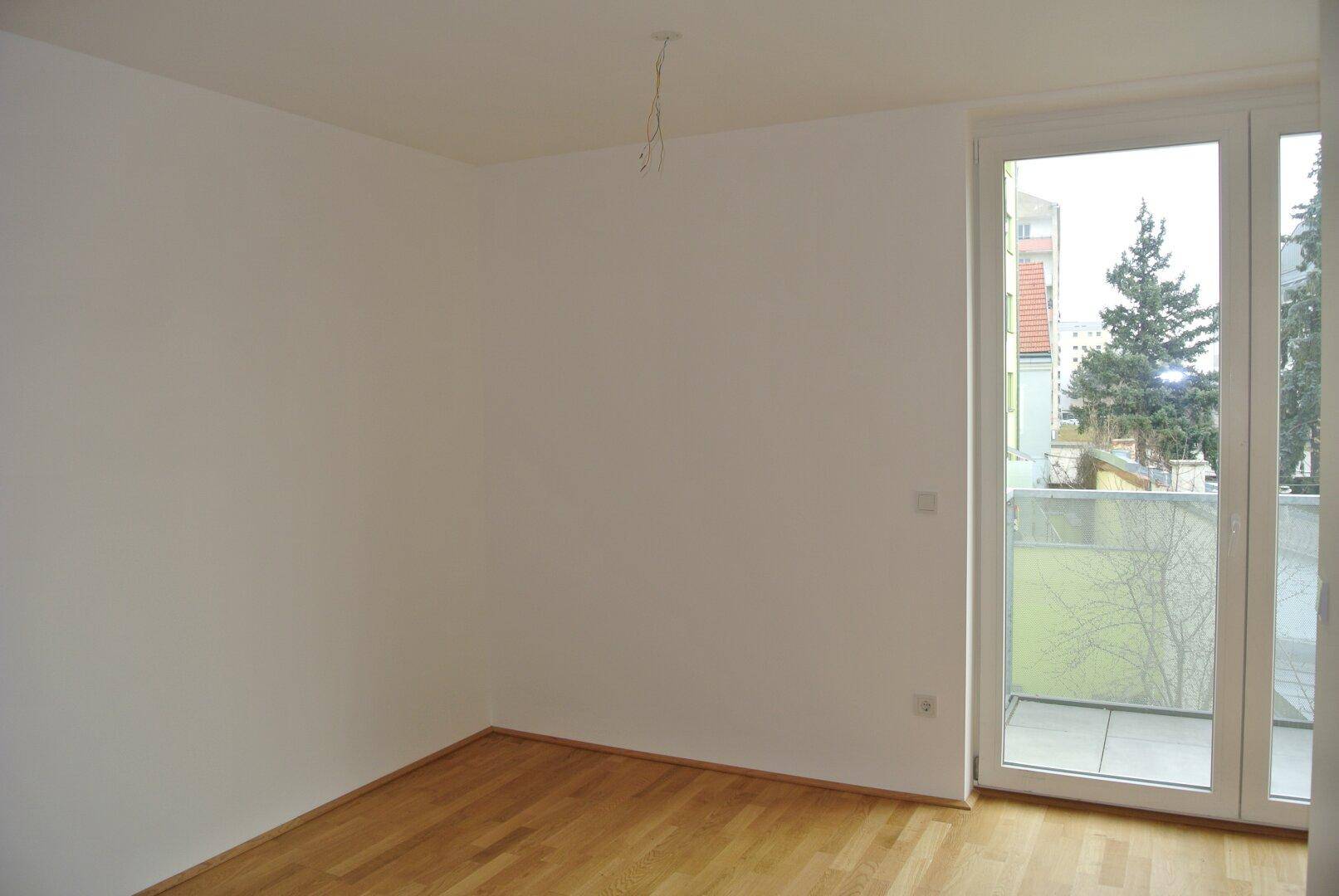 Helles, leeres Zimmer mit Holzboden und Zugang zu einem Balkon, der einen Blick ins Grüne bietet.