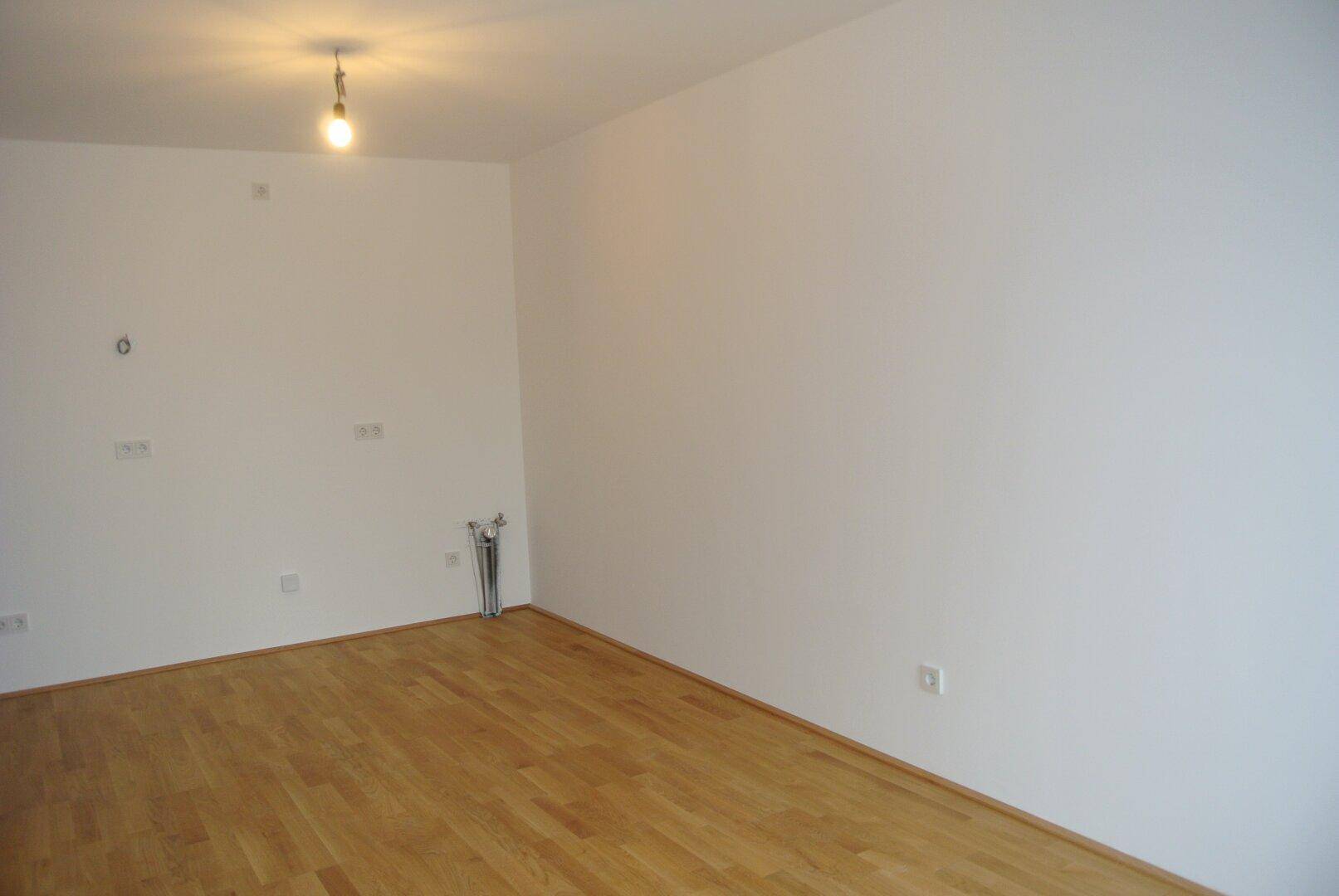 Leeres Zimmer mit hellen Wänden, Parkettboden und sichtbaren Elektroinstallationen.