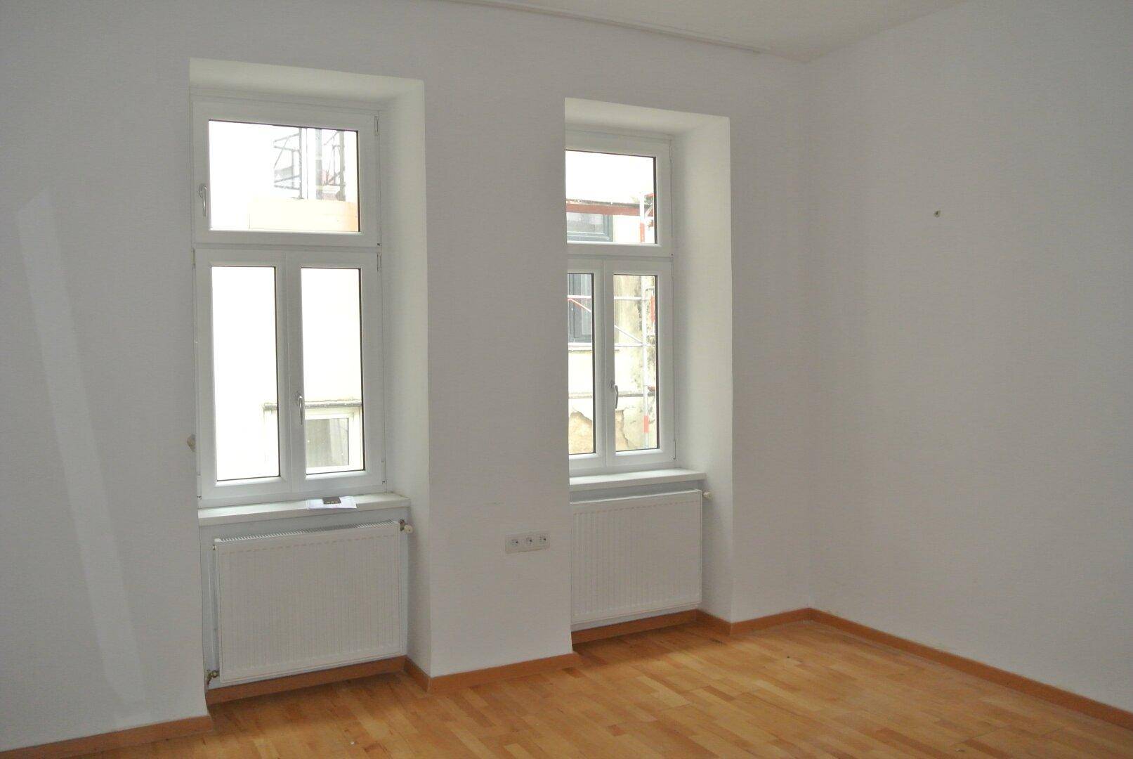 Heller Raum mit zwei Fenstern, Heizkörpern und Holzboden, frisch gestrichen und leerstehend.