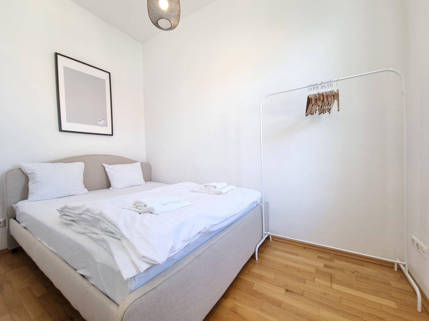 Helles Schlafzimmer mit Holzboden, Doppelbett und einfacher Kleiderstange, minimalistisch gestaltet.