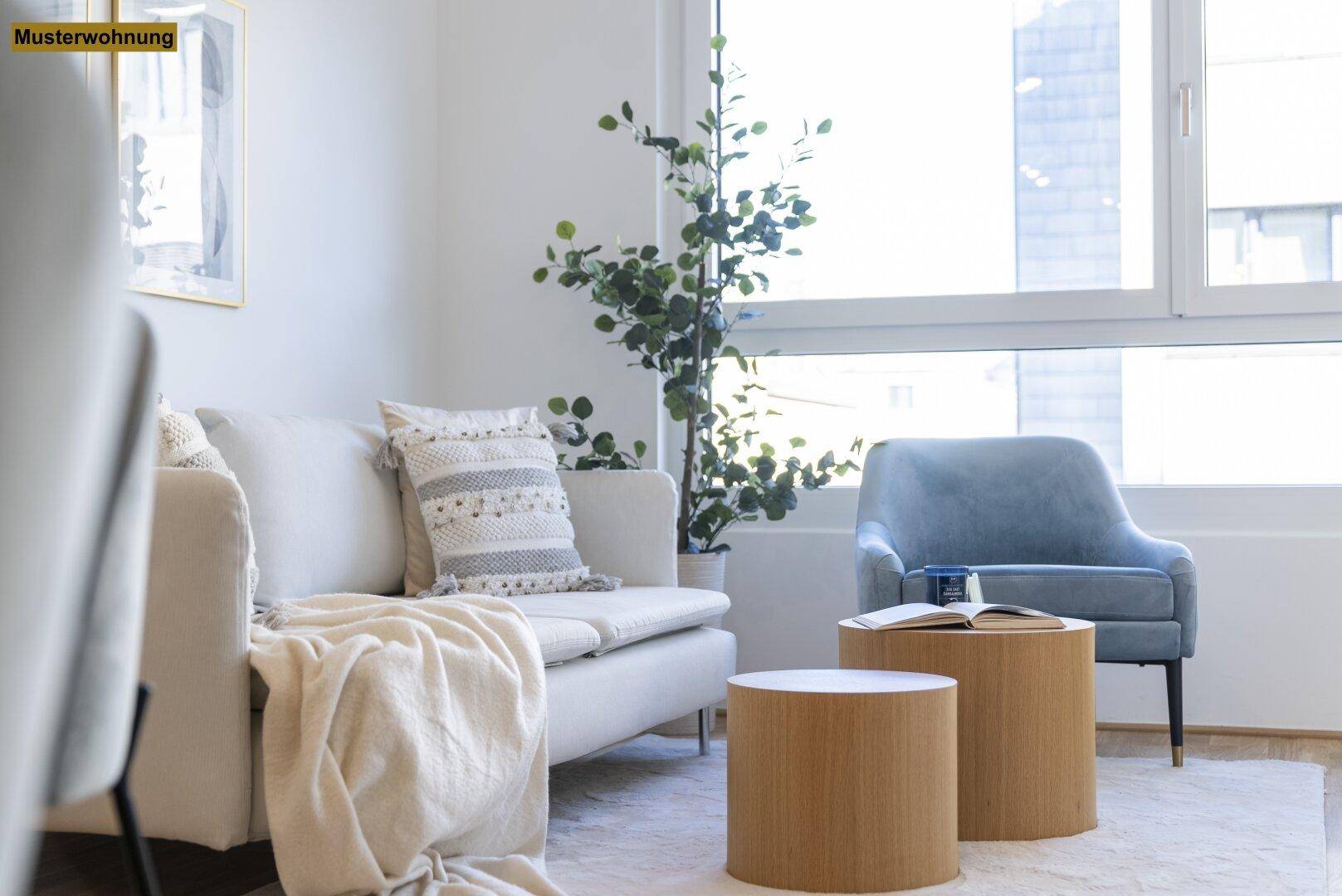 Helles Wohnzimmer mit beigem Sofa, blauem Sessel und großen Fenstern, die viel Tageslicht hereinlassen.