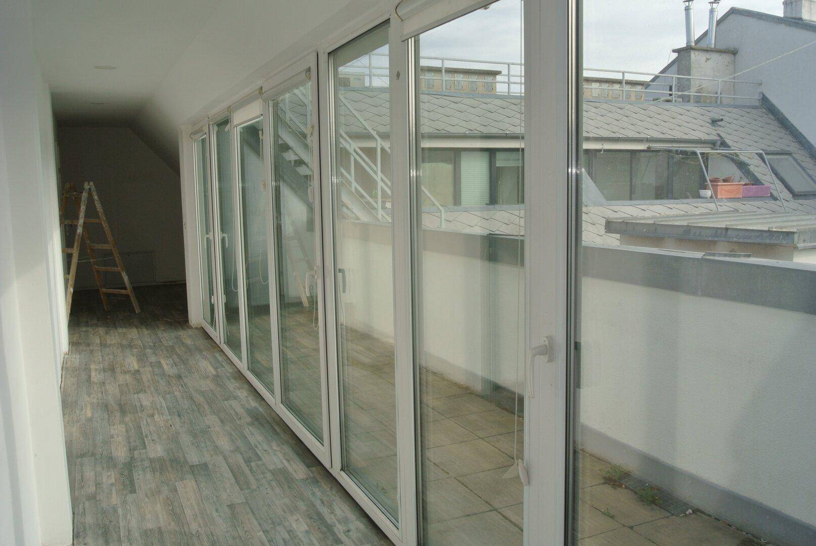Langer Flur mit großen Glasfronten, die zu einem Außenbereich mit Dachblick führen.