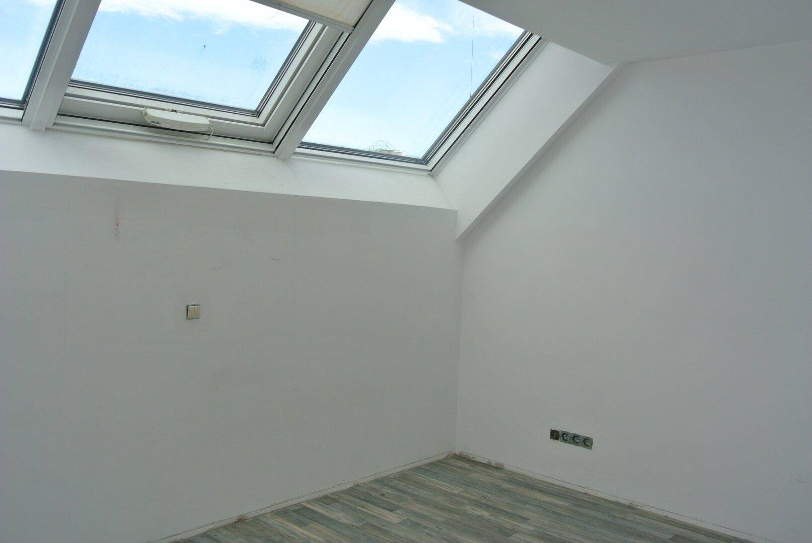 Leerer Raum im Dachgeschoss mit schrägen Wänden, Holzboden und großen Dachfenstern.