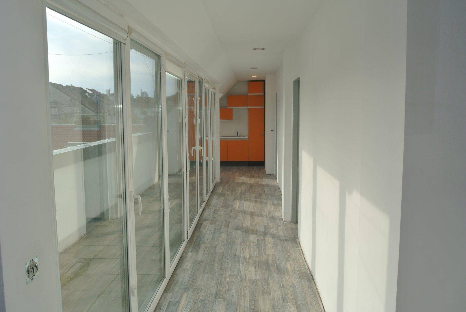 Langer Flur mit Glasfronten und Blick auf eine orangefarbene Küche im Hintergrund.