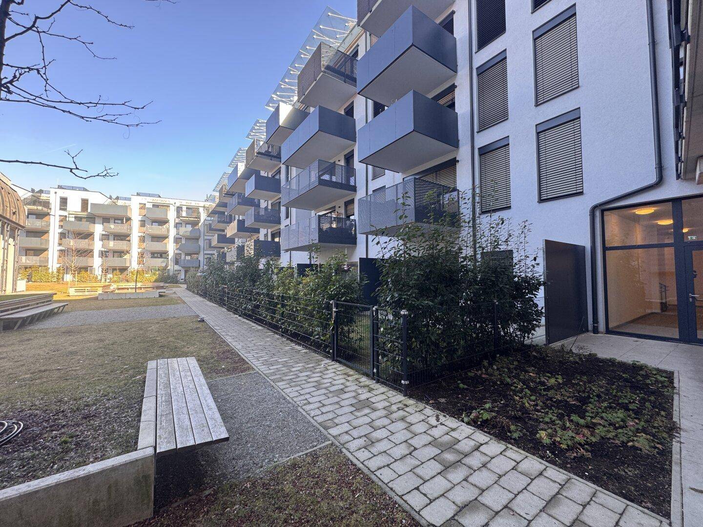 Moderne Wohnanlage mit Balkonen, gepflastertem Weg und Sitzgelegenheiten im Gemeinschaftsgarten.
