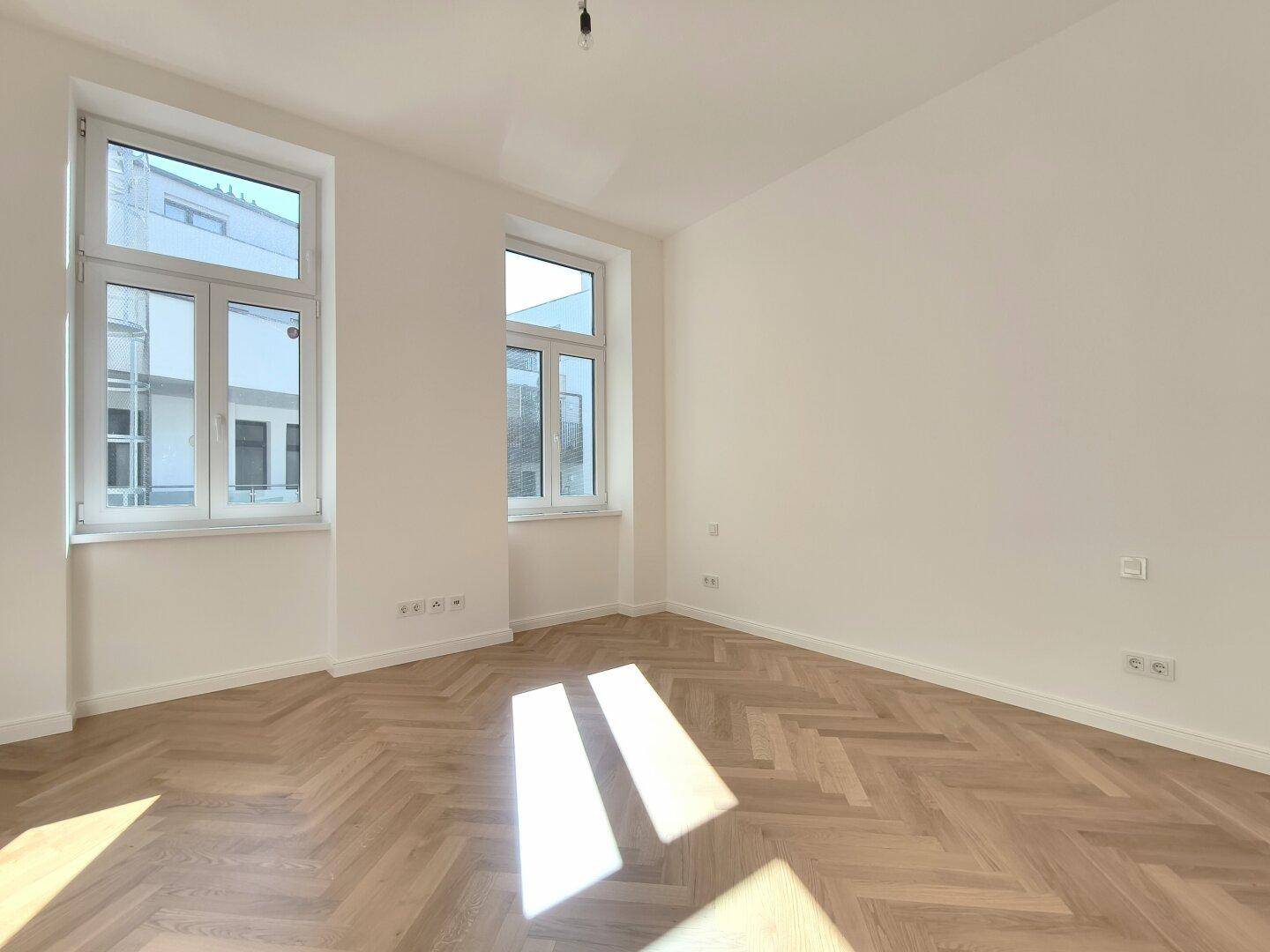 Lichtdurchflutetes Zimmer mit Fischgrätparkett und großen Fenstern, die viel Tageslicht hereinlassen.