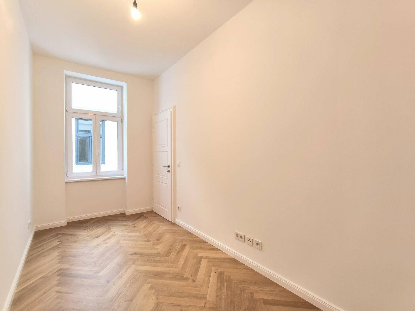 Gemütliches Zimmer mit Fischgrätparkett und einem Fenster, bietet direkten Zugang zu weiteren Räumen.