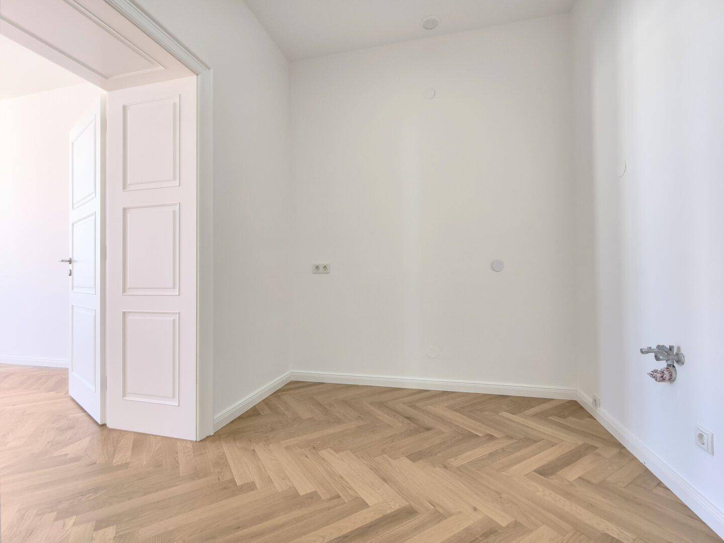 Leerer Raum mit Fischgrätparkett und Anschlüssen für eine Küche, bereit für die Installation.