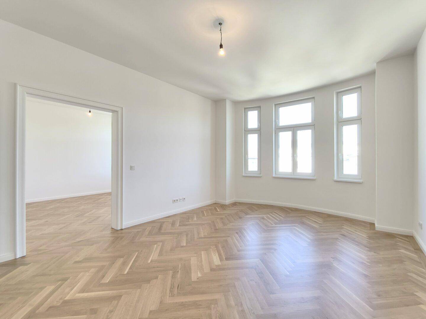 Helles Zimmer mit elegantem Fischgrätparkett und mehreren Fenstern, ideal für Wohn- oder Arbeitsbereich.