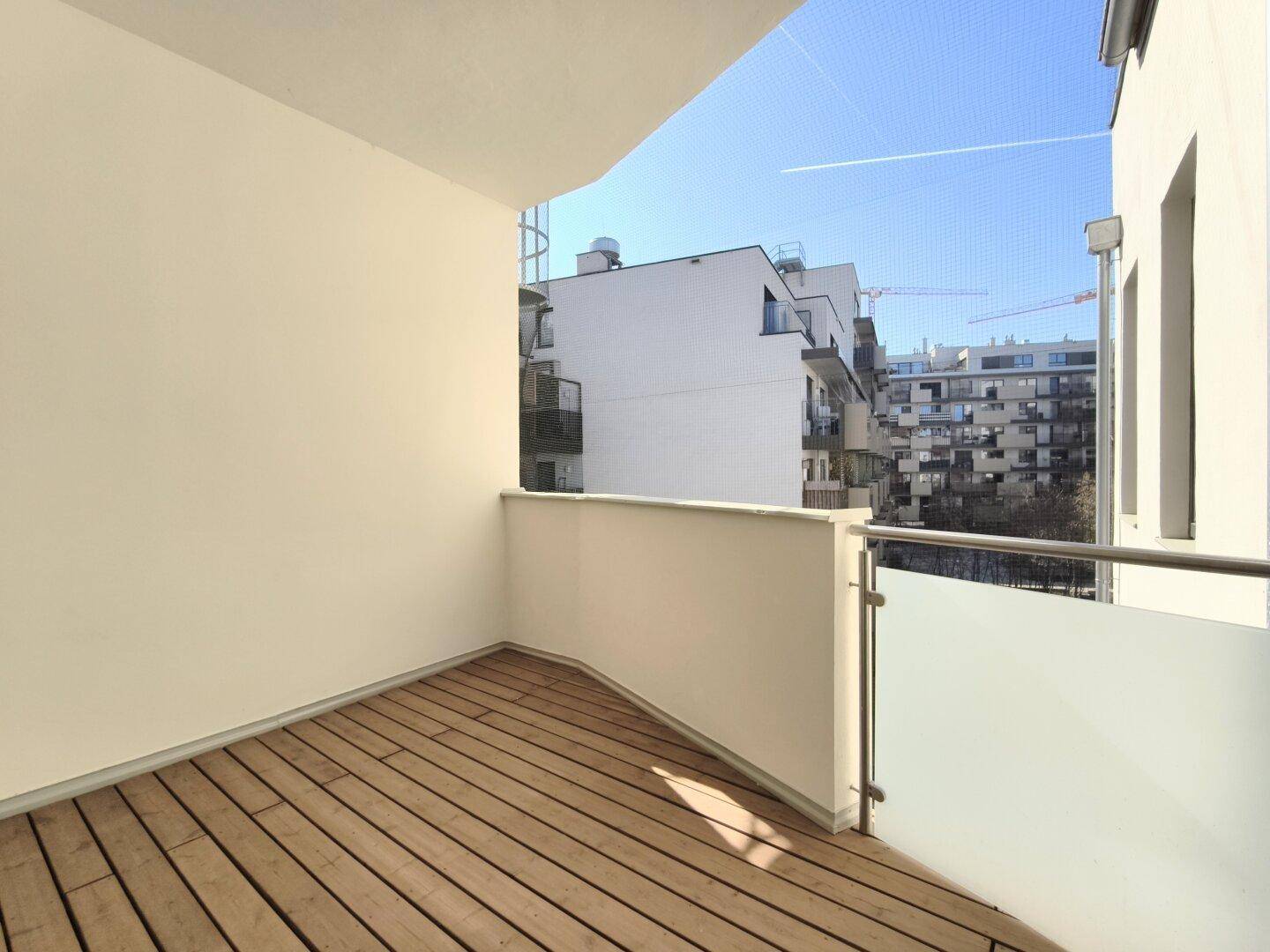 Sonniger Balkon mit Holzdielen und modernem Glasgeländer, umgeben von städtischer Architektur.