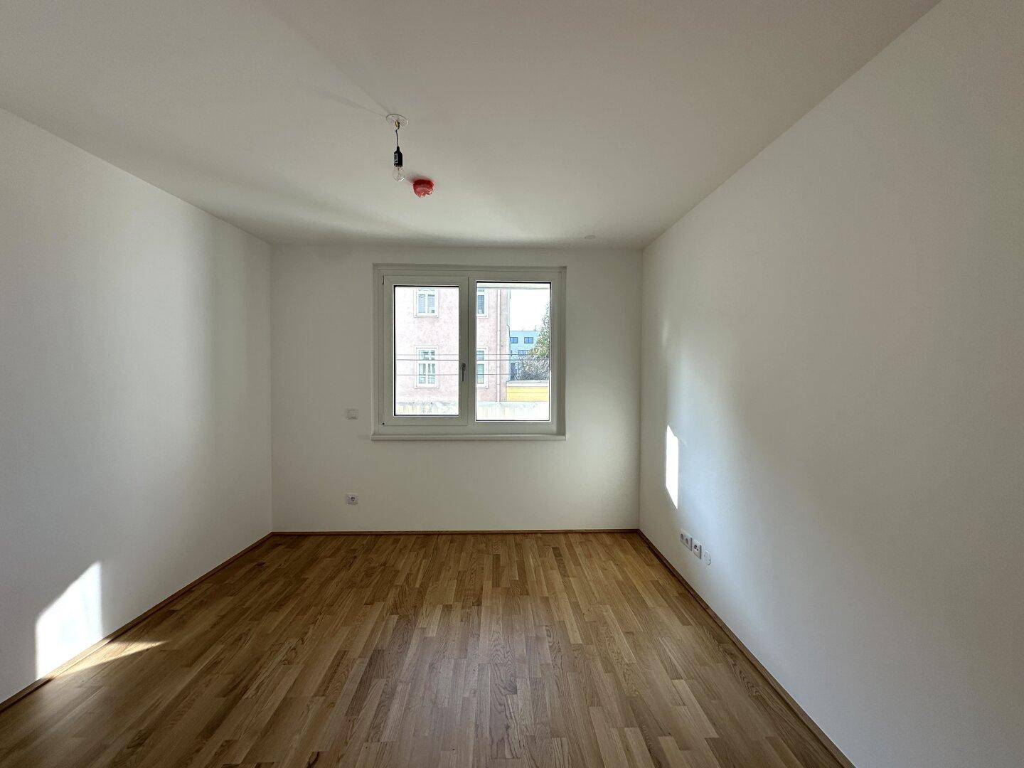 Helles Schlafzimmer mit Parkettboden und großem Fenster, das den Raum erhellt.
