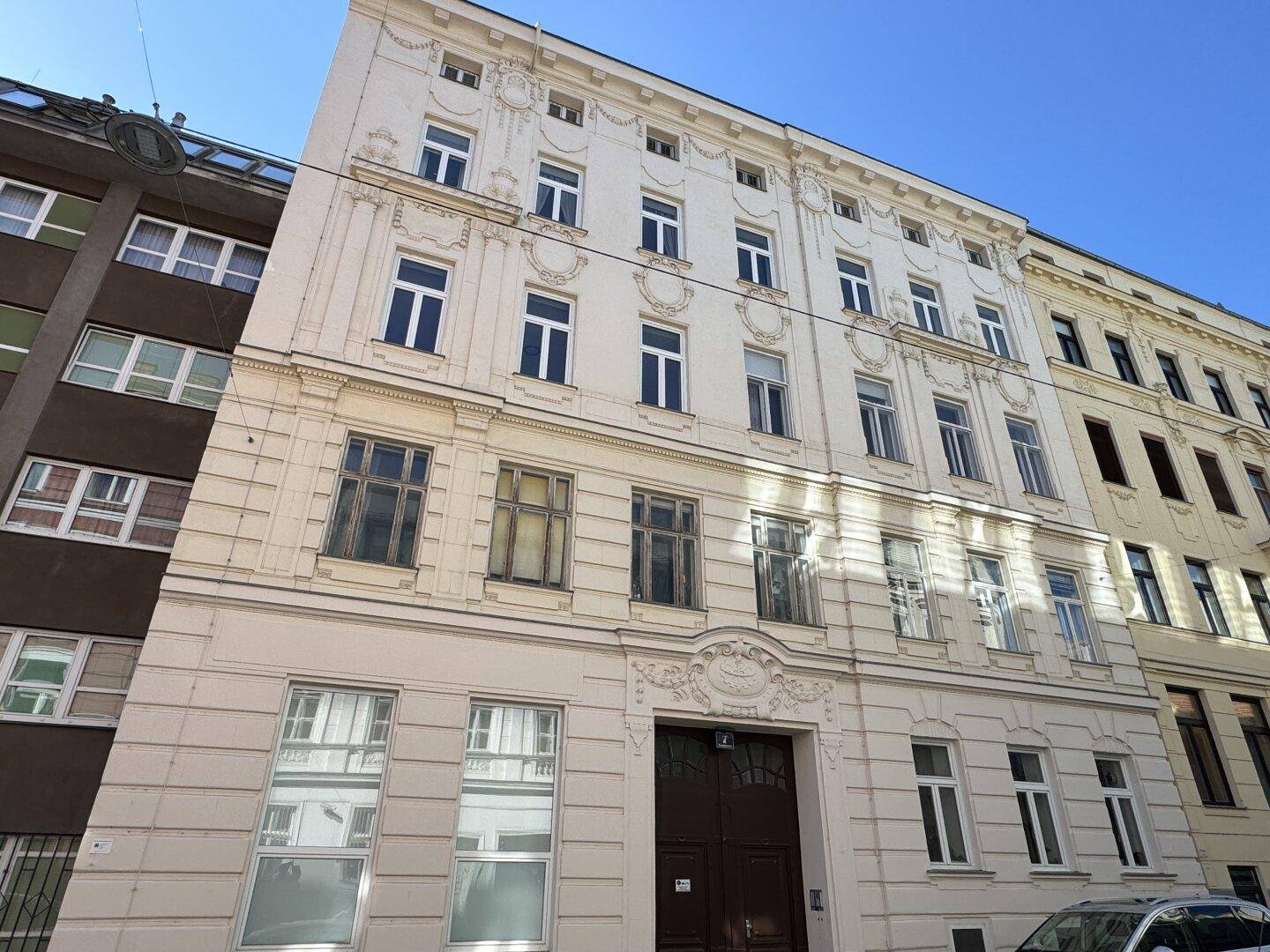 Historische Fassade eines Altbaus mit dekorativen Details und angrenzenden Gebäuden unter blauem Himmel.
