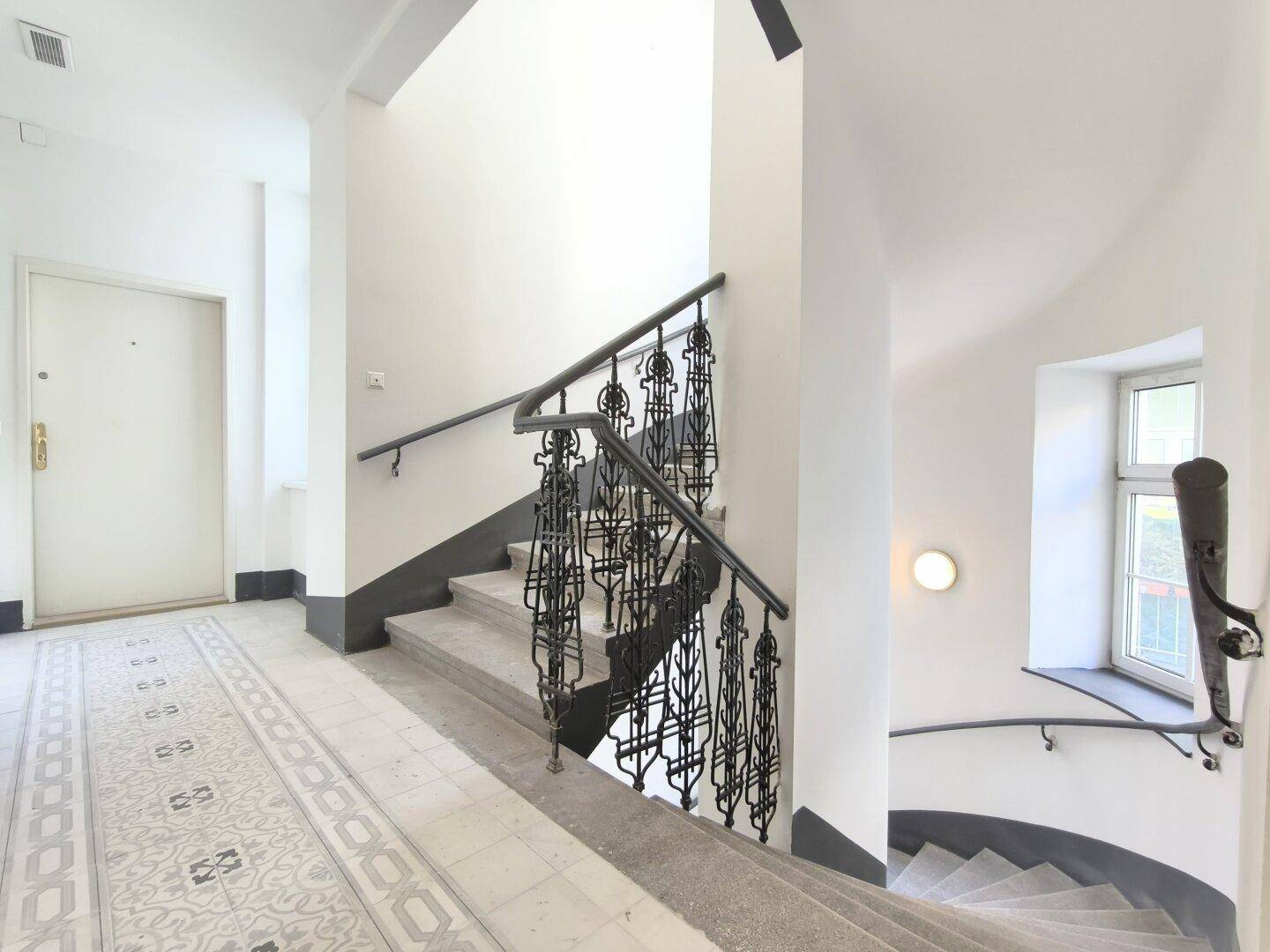 Elegantes Treppenhaus mit schmiedeeisernem Geländer und Tageslicht durch ein Fenster.