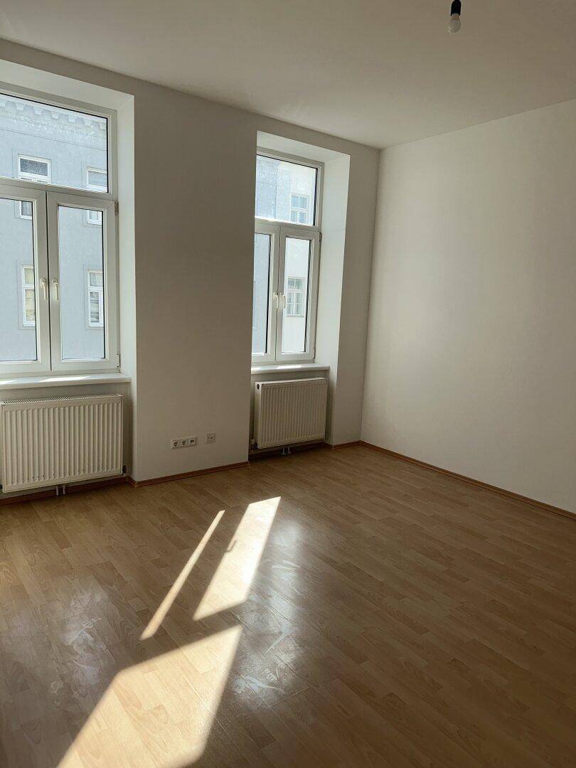 Heller, leerer Raum mit Holzparkettboden, weißen Wänden und zwei großen Fenstern.