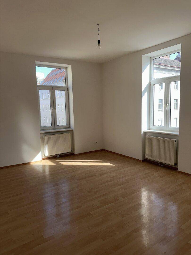 Heller, leerer Raum mit Holzparkettboden, weißen Wänden und zwei großen Fenstern.