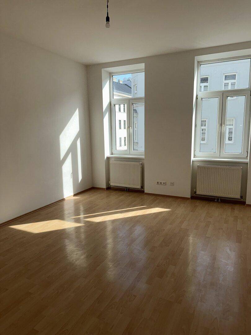 Lichtdurchfluteter, leerer Raum mit Holzparkettboden, weißen Wänden und zwei großen Fenstern.