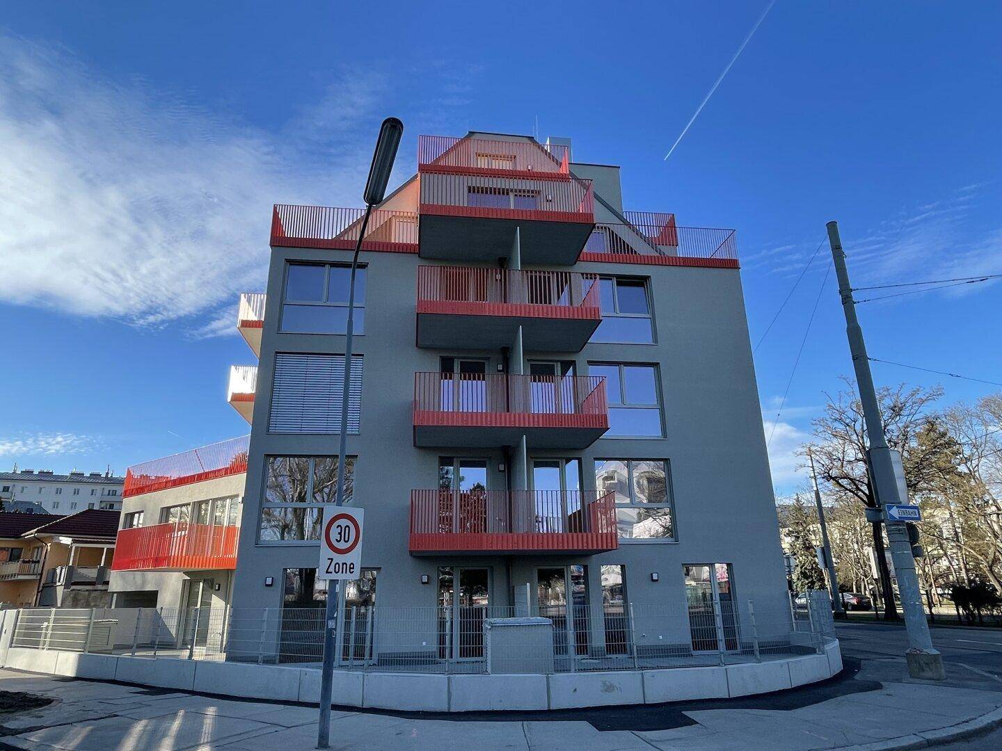 Neubau-Wohngebäude mit markanten roten Balkonen und großen Fenstern in urbaner Umgebung.