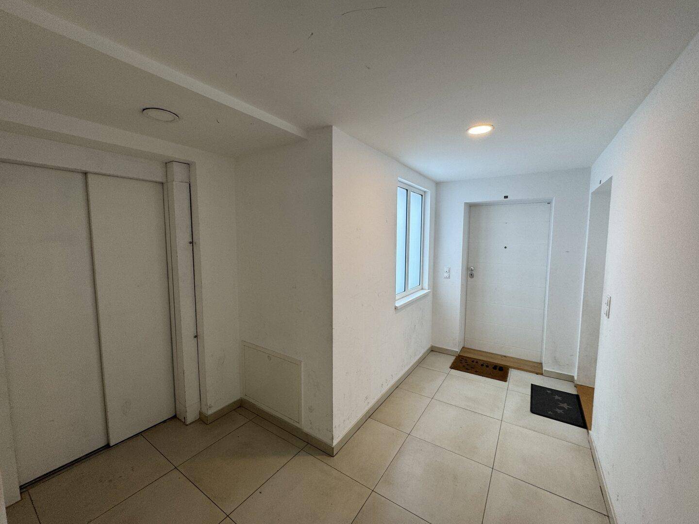 Eingangsbereich mit hellen Fliesen, weiß gestrichenen Wänden, einem Fenster und einer Aufzugstür.
