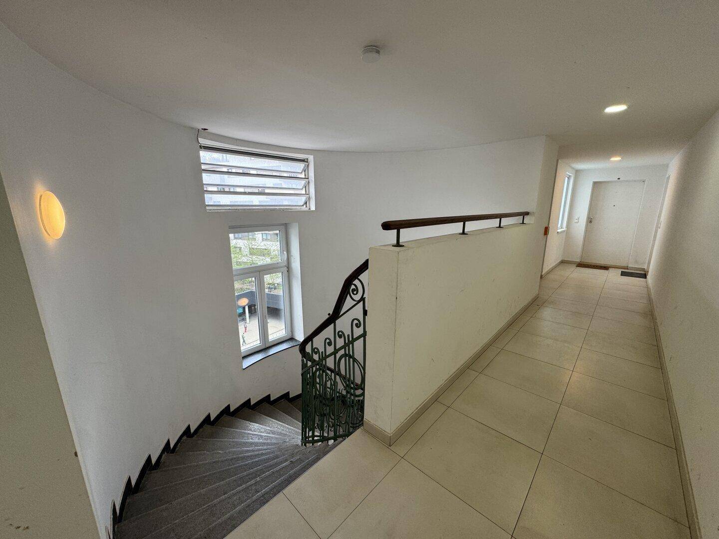 Treppenhaus mit geschwungener Treppe, schmiedeeisernem Geländer und großen Fenstern für natürliches Licht.