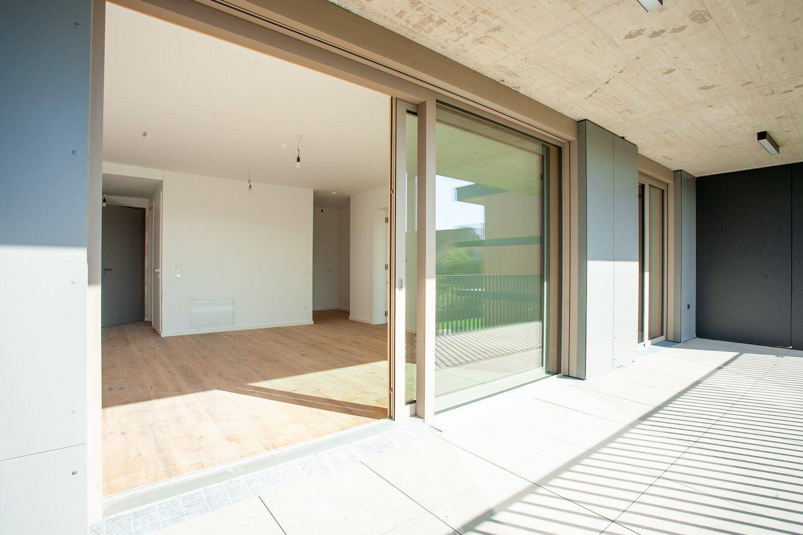 Helle, leere Wohnfläche mit Holzboden und großen Schiebetüren, die auf eine Terrasse führen.