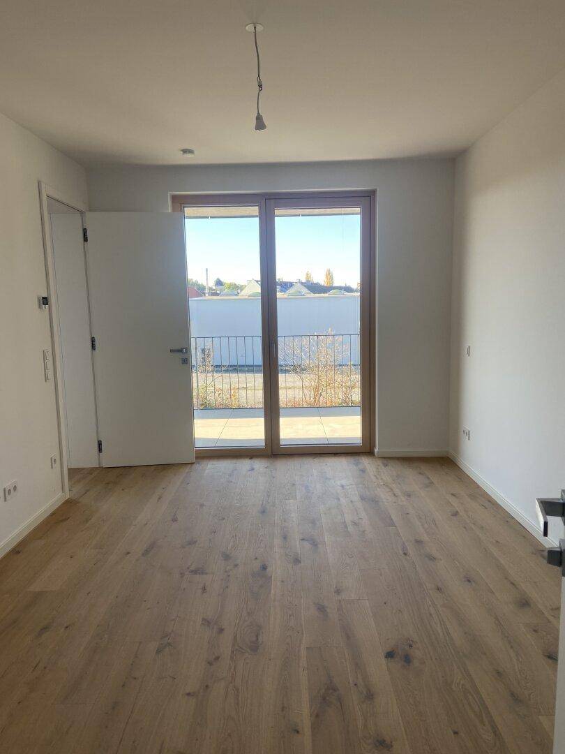 Leeres Zimmer mit Holzboden und weiß gestrichenen Wänden, mit Zugang zu einem Balkon.