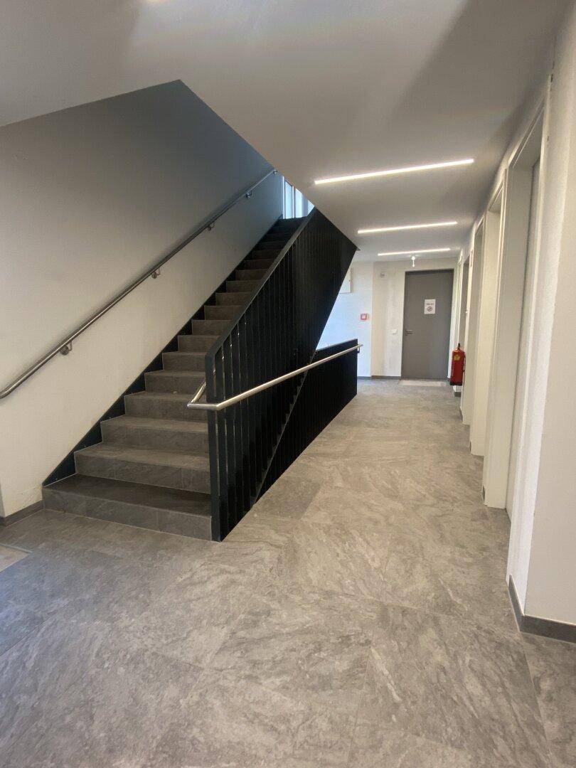 Moderner Treppenaufgang und Flur in einem Mehrfamilienhaus mit grauen Fliesen und Handläufen.