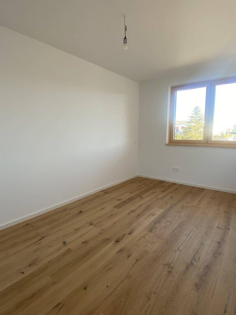 Helles, leeres Zimmer mit Holzboden und einem Fenster, das viel Tageslicht hereinlässt.
