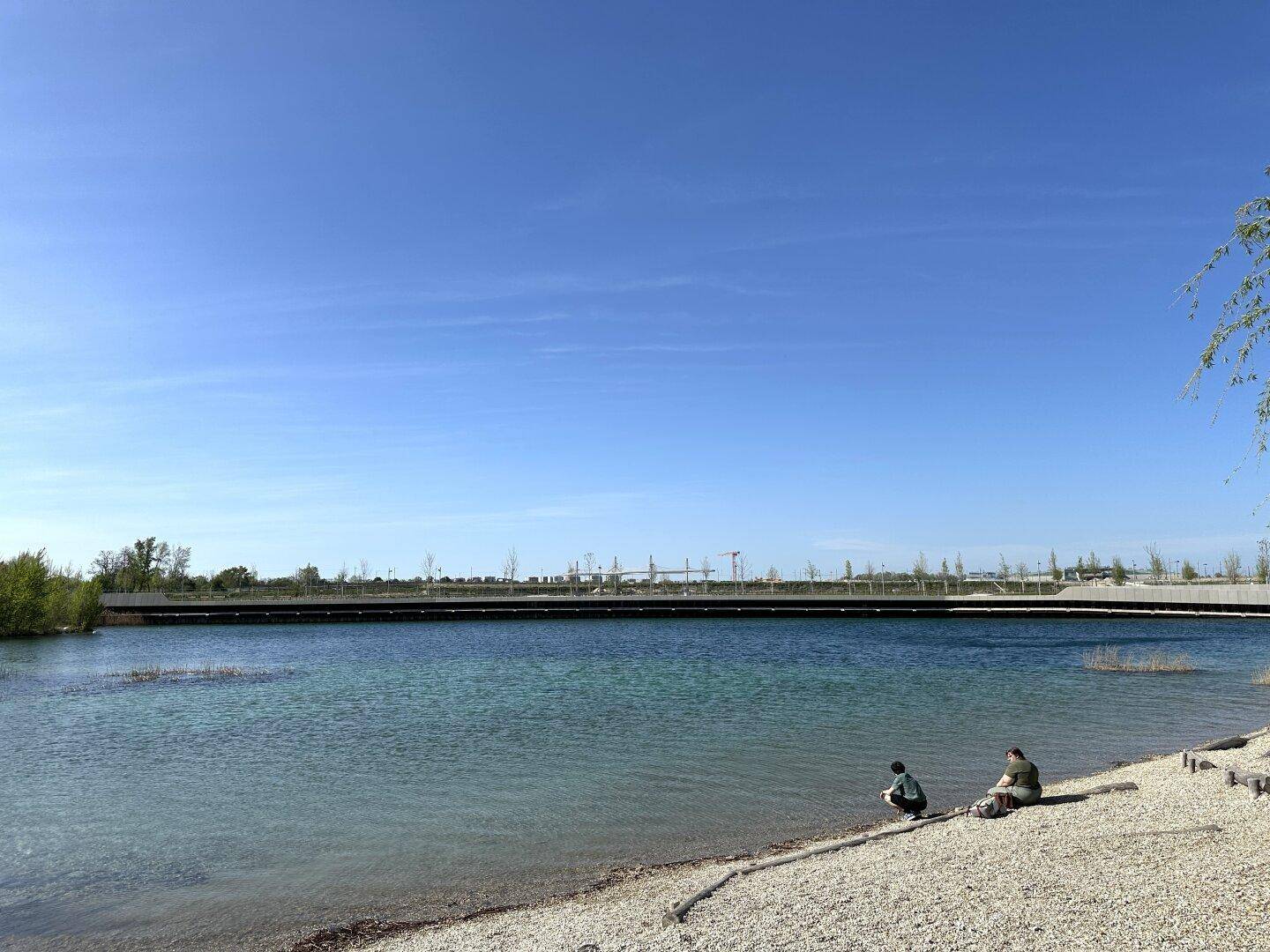 Ruhiger Seeuferbereich mit Kiesstrand und Blick auf die weite Landschaft unter blauem Himmel.