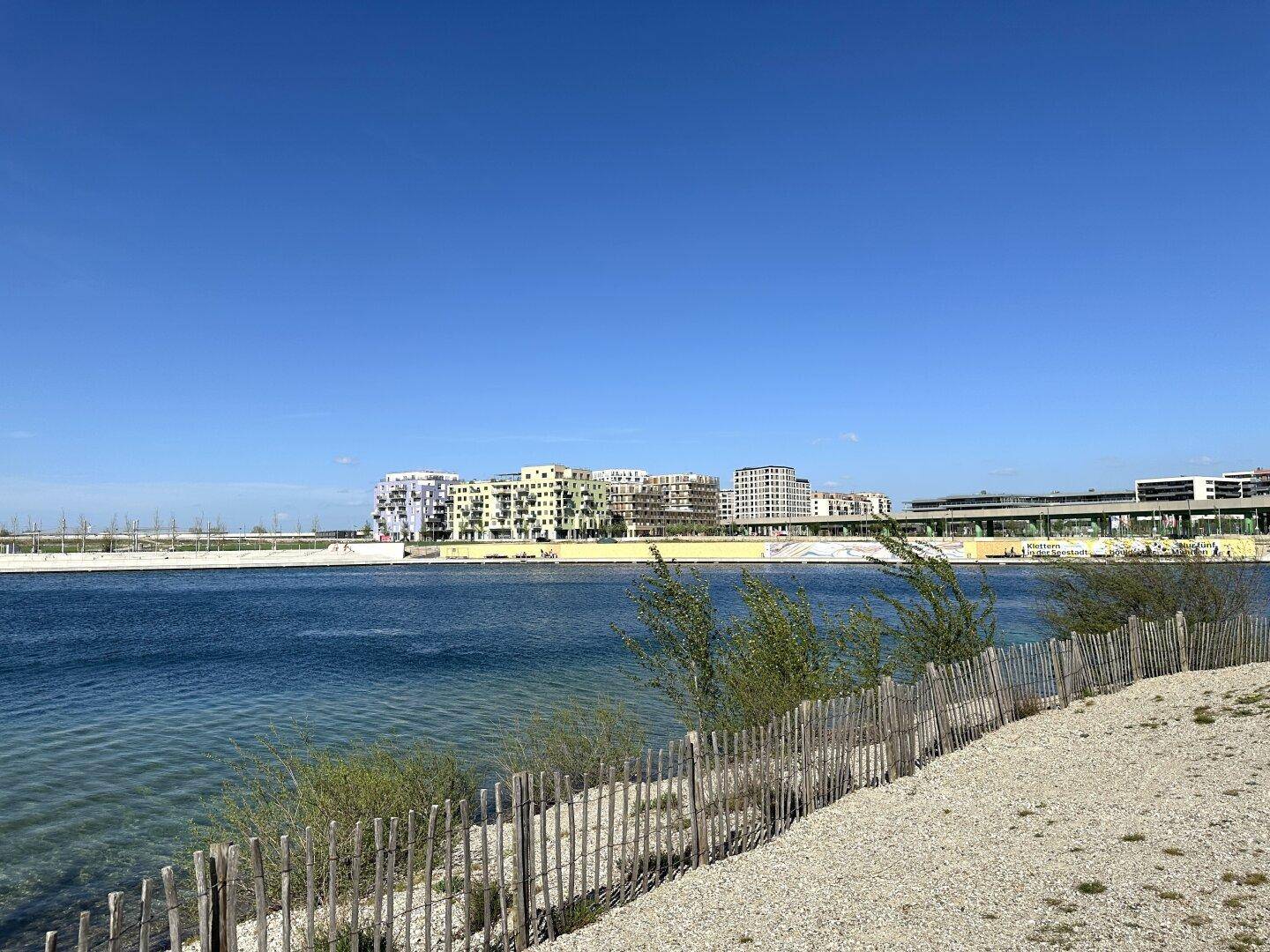 Weitläufiger See mit Uferbefestigung und Blick auf moderne Gebäude am Horizont.