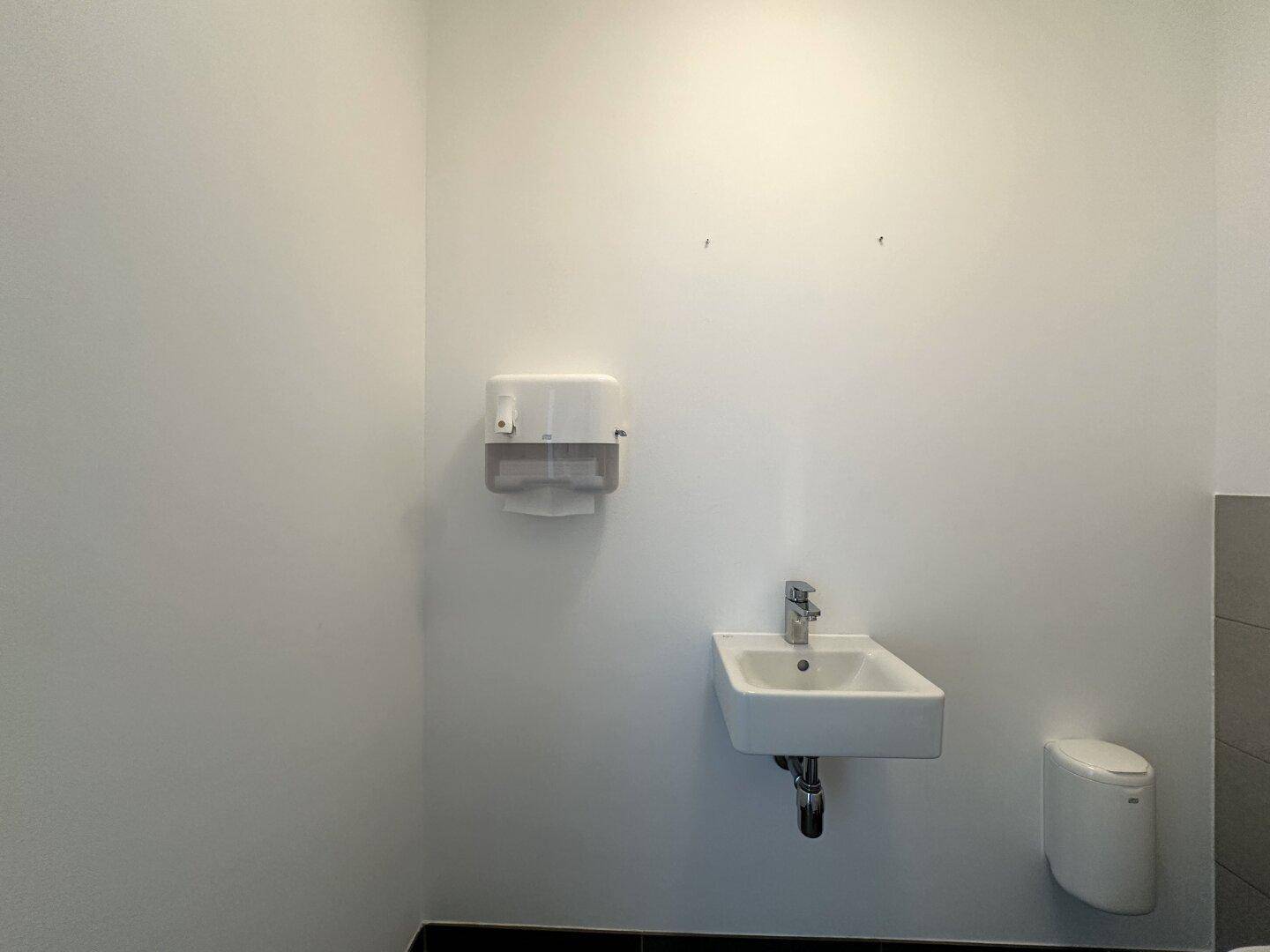 Helles Gäste-WC mit kleinem Waschbecken, Seifenspender und dunklem Fliesenboden.