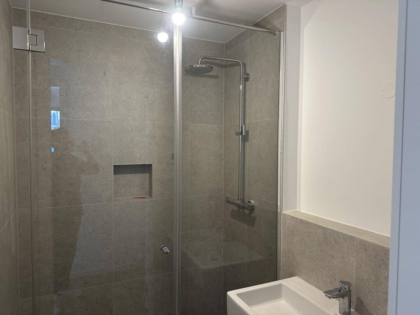 Modernes Badezimmer mit begehbarer Dusche, Glasabtrennung und grauen Wandfliesen.