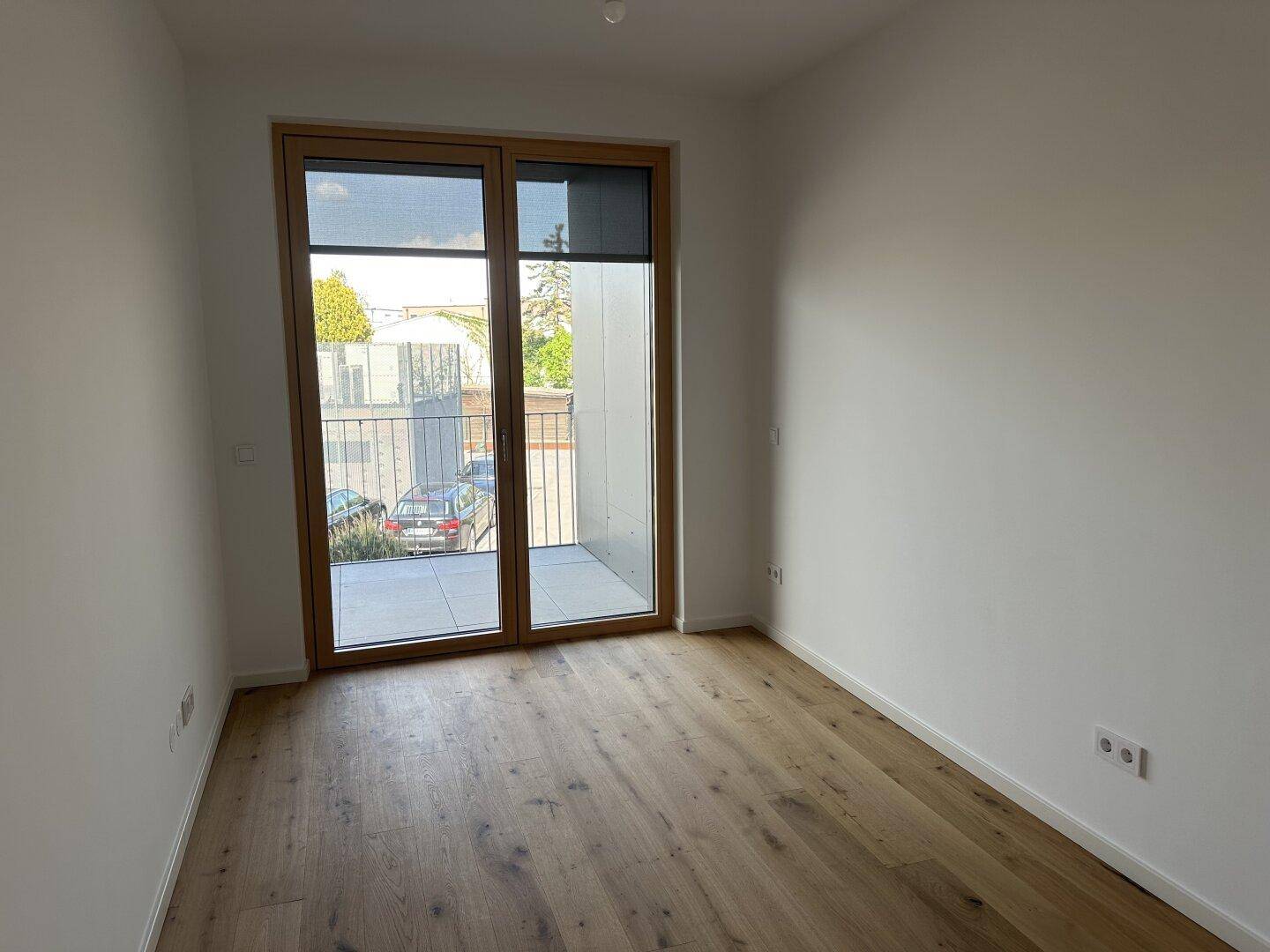 Leeres Zimmer mit Eichenparkettboden und bodentiefen Fenstern, die zu einem Balkon führen.