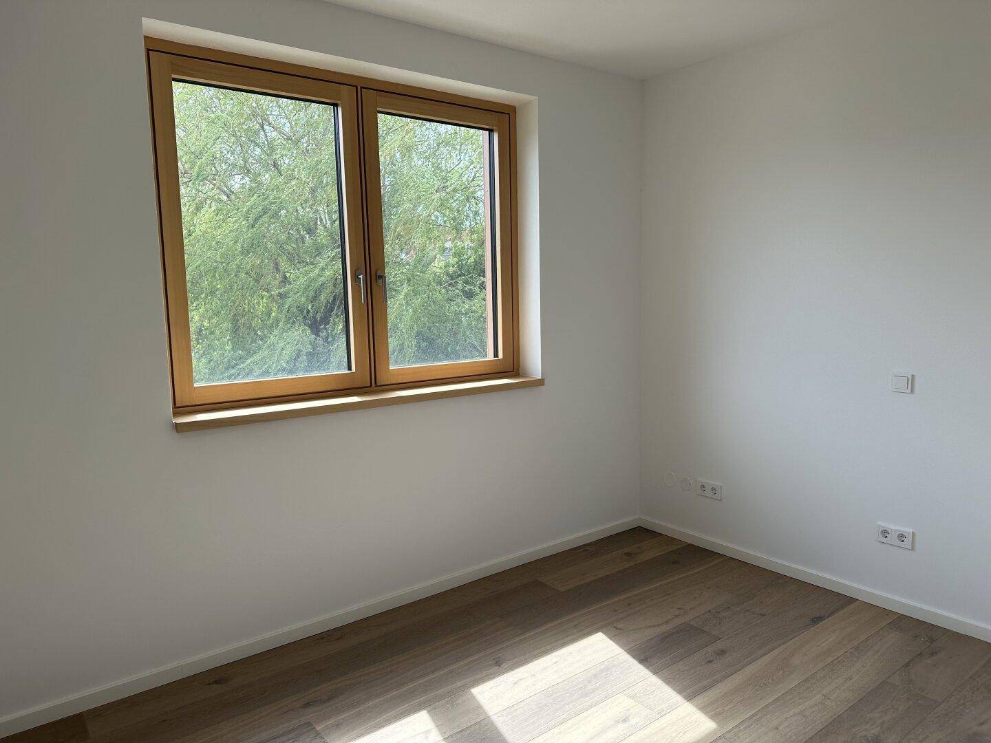 Helles Schlafzimmer mit Holzplankenboden und einem Fenster mit Blick ins Grüne.