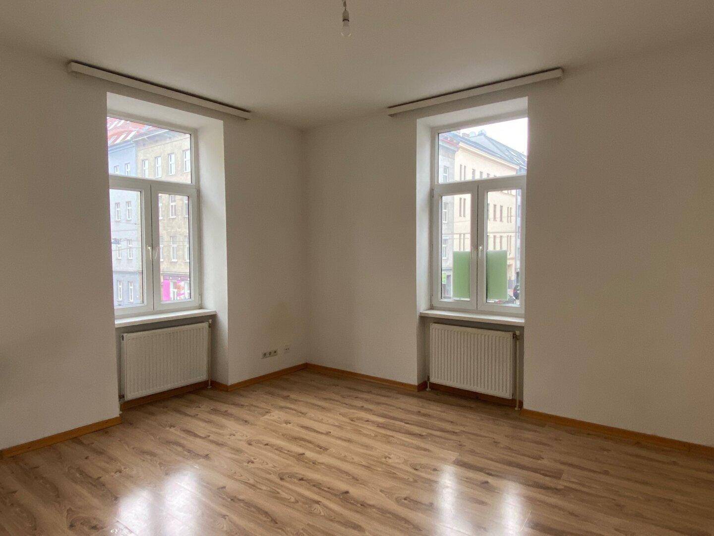Heller, leerer Raum mit Laminatboden und zwei Fenstern zur Straße.
