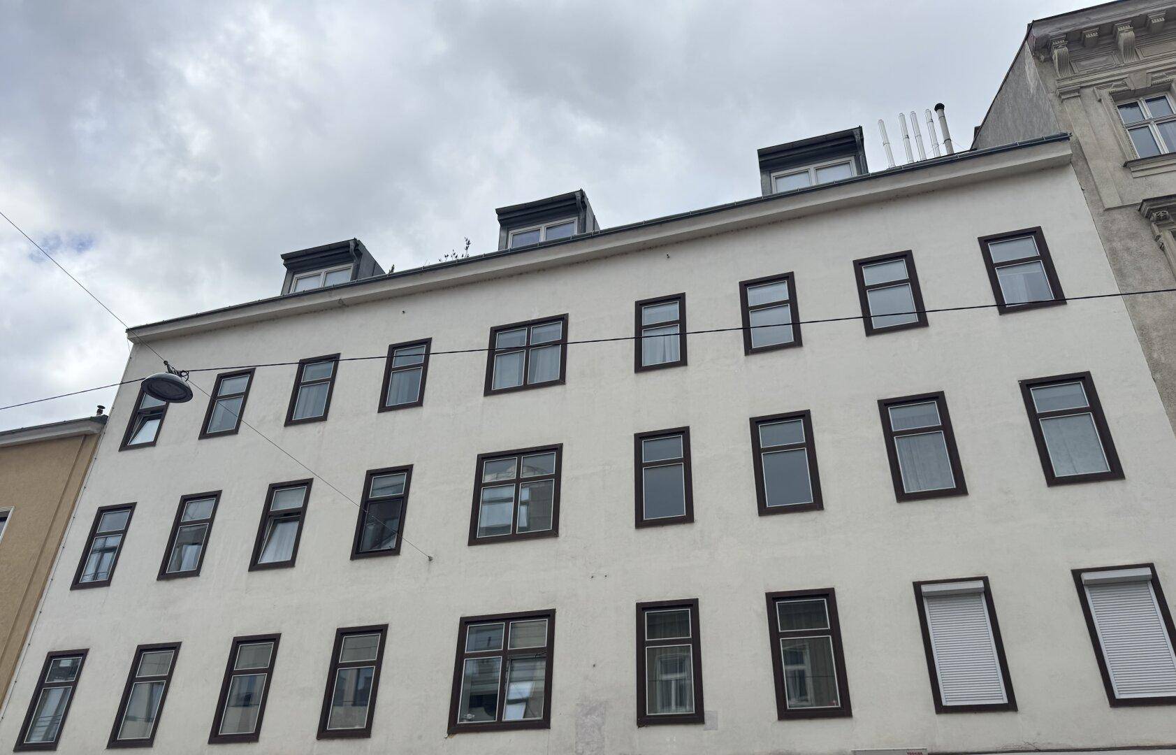 Ansicht eines mehrstöckigen Gebäudes mit weißer Fassade, vielen Fenstern und Dachgauben unter bewölktem Himmel.