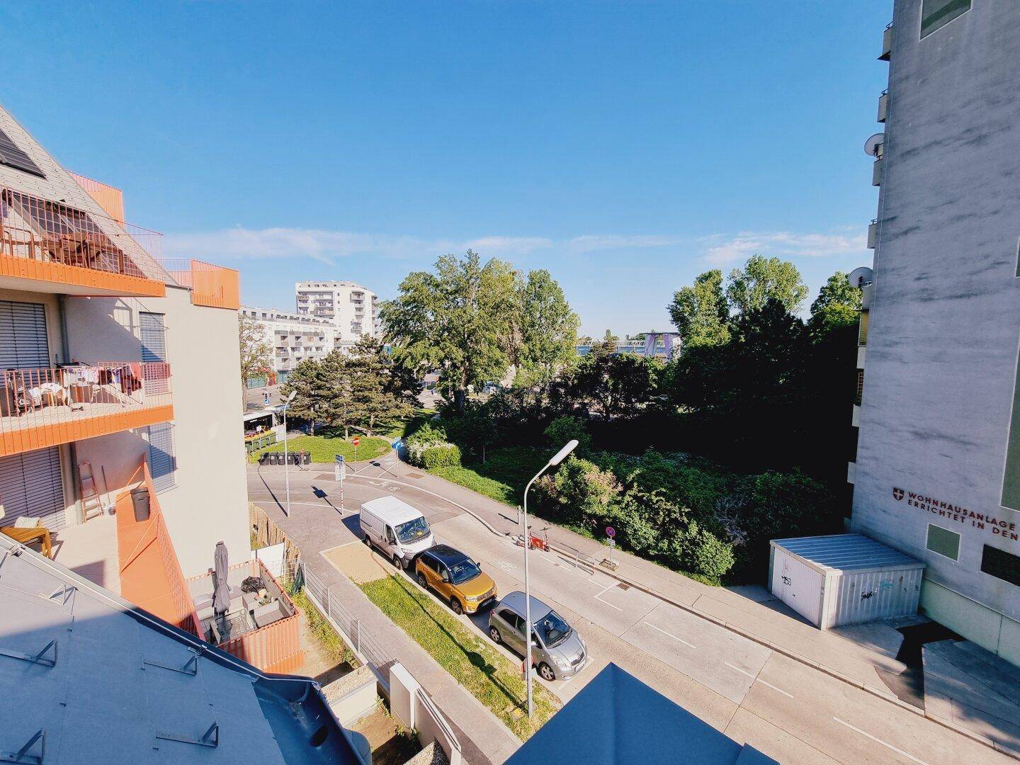 Blick von oben auf die Straße, umliegende Gebäude und viel Grün unter blauem Himmel.