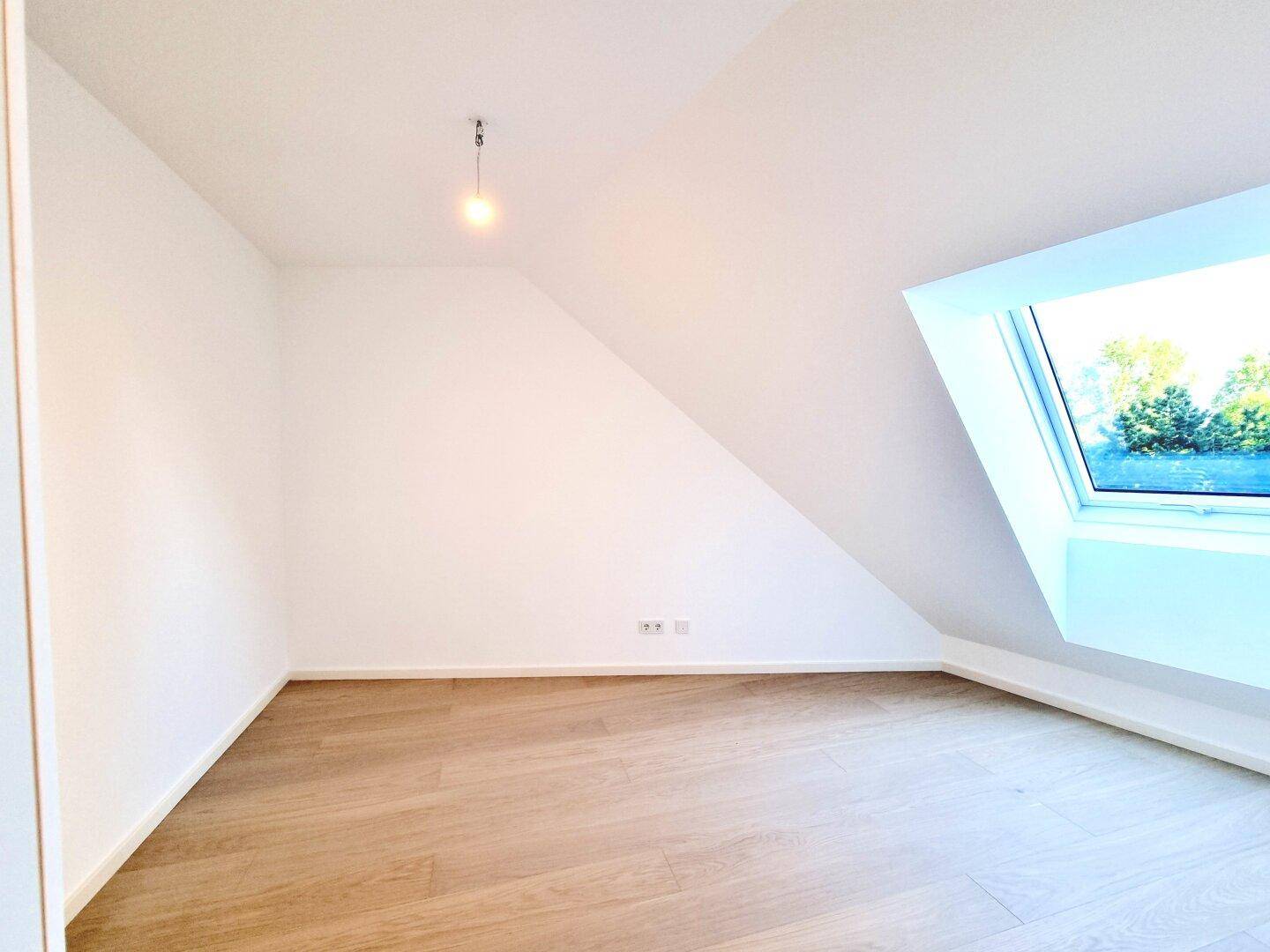 Helles Dachgeschosszimmer mit Holzboden und großem Dachfenster, das einen Blick ins Grüne bietet.