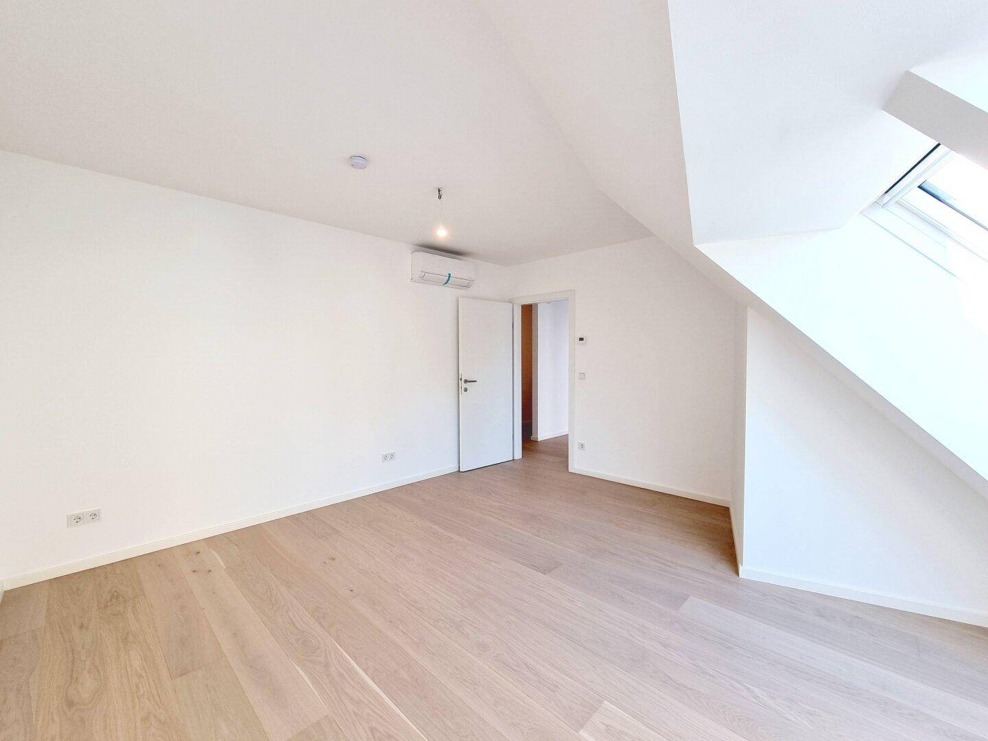 Helles Dachgeschosszimmer mit Holzboden, Klimaanlage und Zugang zu einem weiteren Raum.