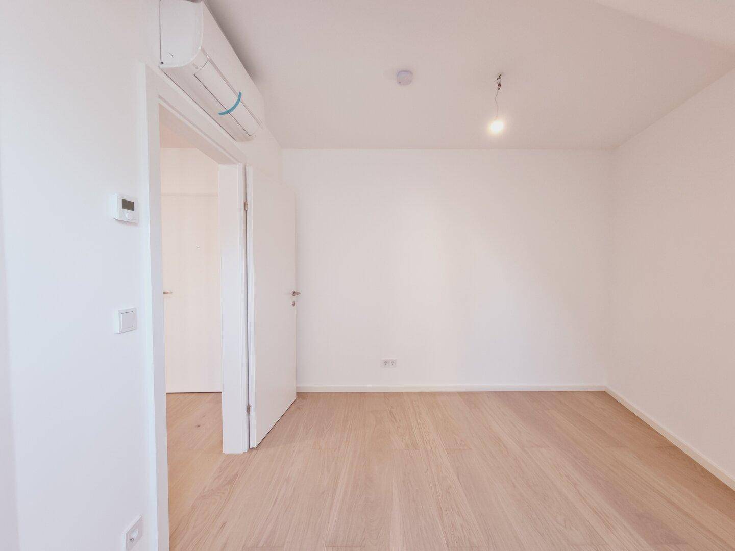 Leeres Zimmer mit Holzboden, Klimaanlage und weiß gestrichenen Wänden, bereit zur Einrichtung.