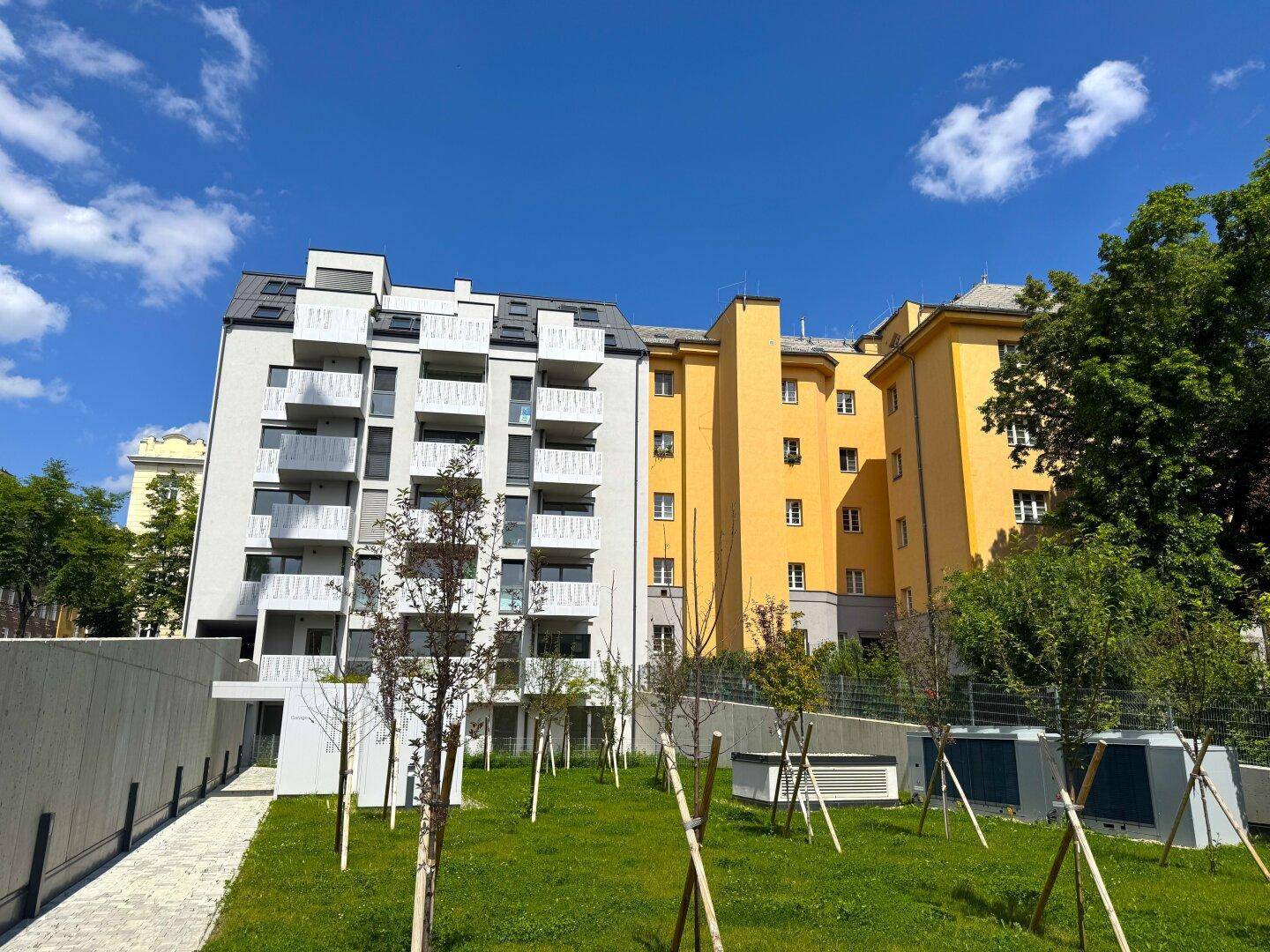 Zeitgenössisches Wohngebäude mit weißer Fassade neben einem älteren gelben Gebäude, umgeben von Grünflächen.