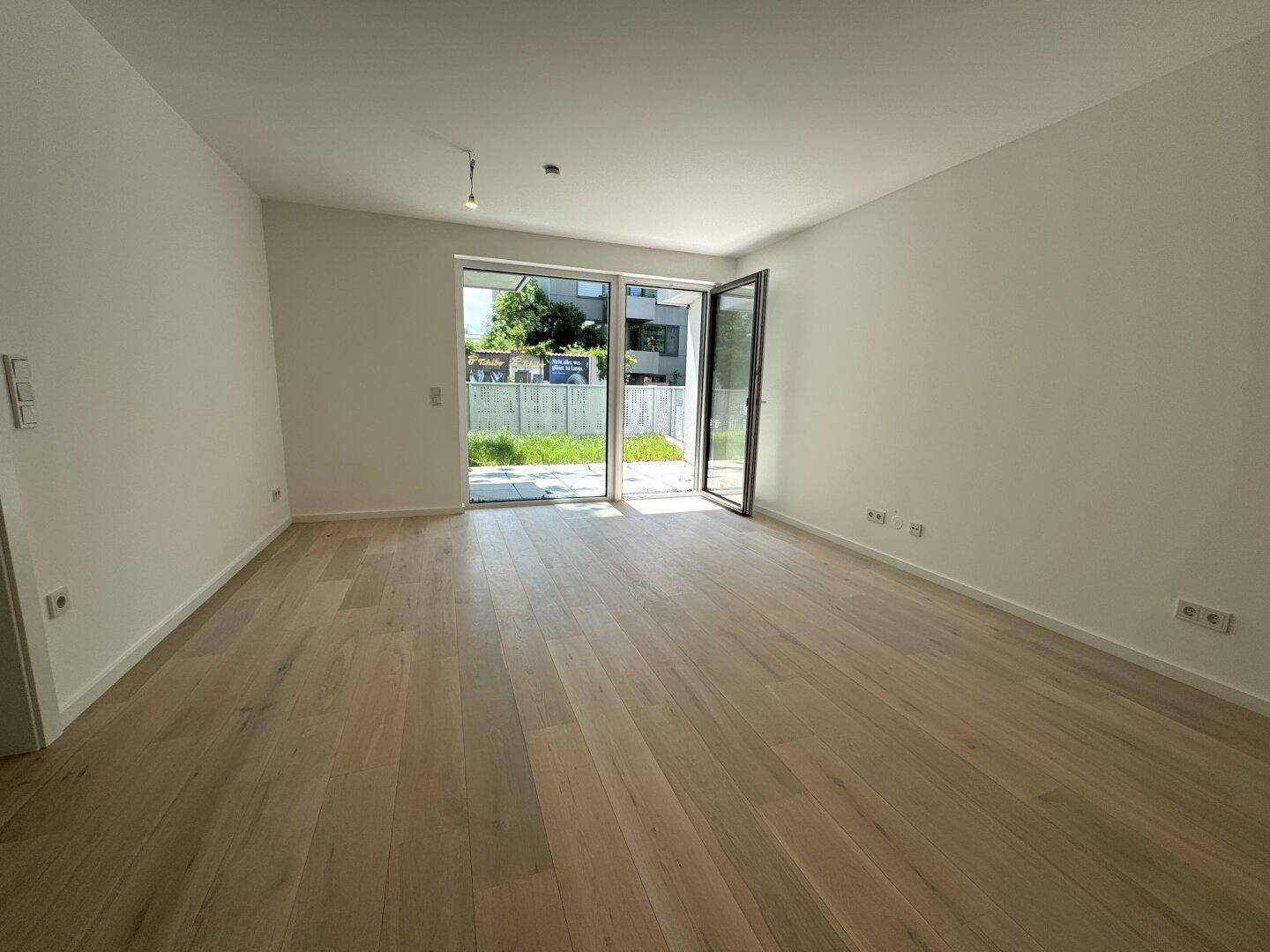 Geräumiger, heller Wohnraum mit Holzboden und offener Glastür, die viel Licht hereinlässt.